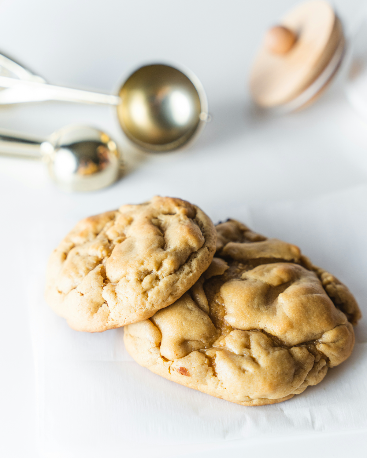 Two thick golden cookies on parchment with cookie scoops behind